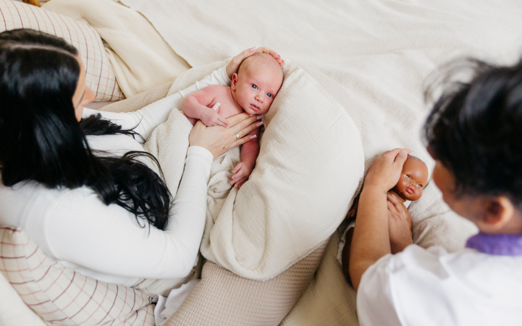 Babymassage met moeder en baby in bed naast kraamverzorgende met een pop. Alle kraamverzorgenden van Naviva zijn opgeleid in babymassage.
