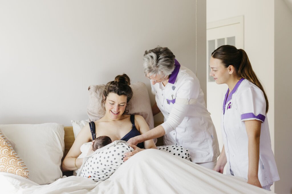 Moeder geeft borstvoeding in bed aan baby. Twee kraamverzorgende staan naast het bed. Leren en werken bij Naviva Kraamzorg.